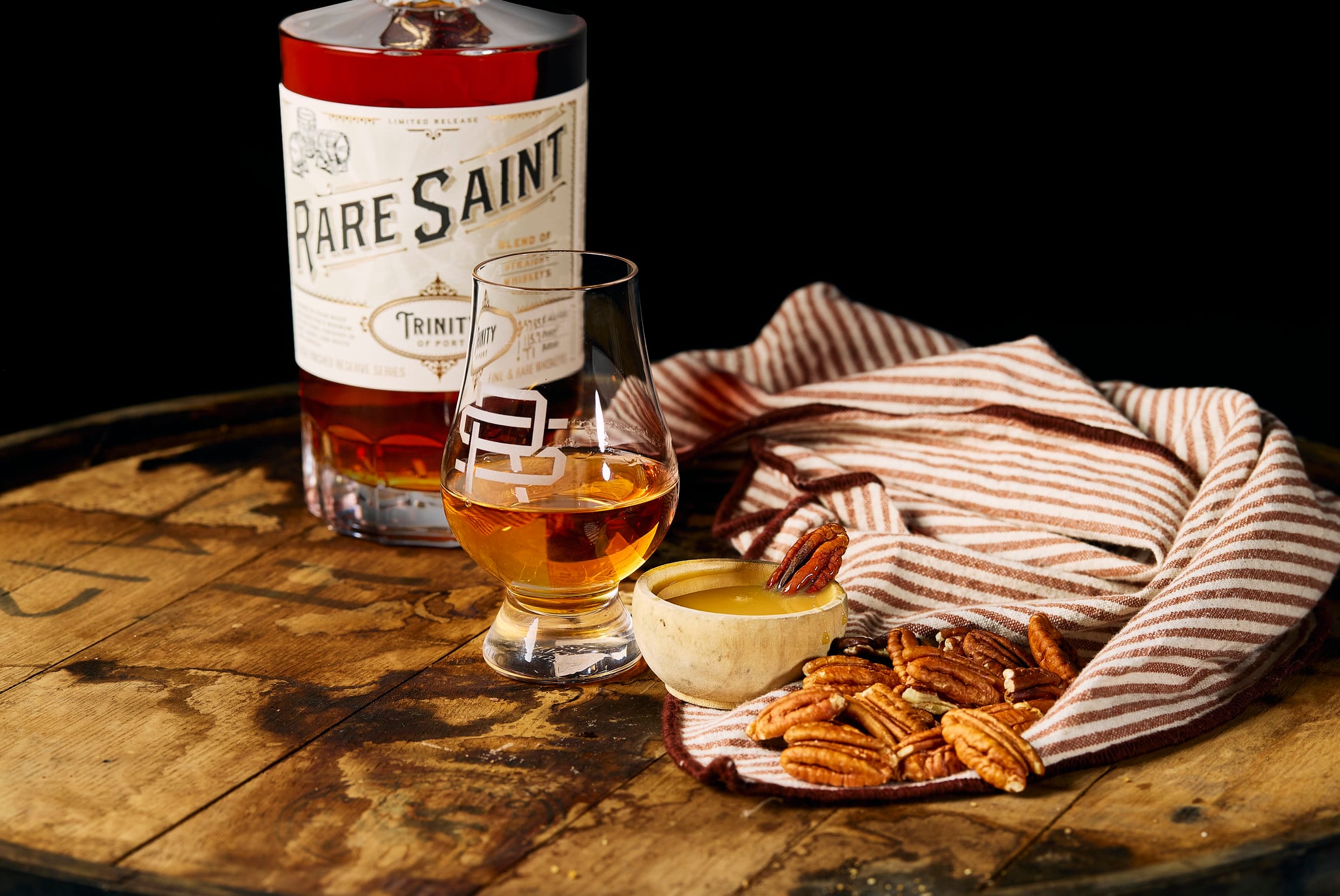 A bottle of Rare Saint Trinity of Port whiskey on a wooden barrel beside a whiskey glass, pecans, and a small bowl with caramel sauce, draped in a striped cloth.